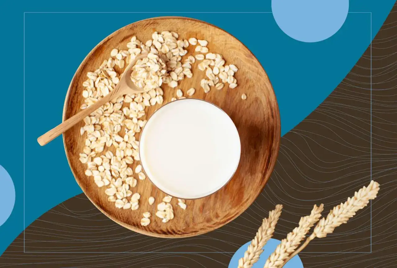 Ein Glas cremiger Hafermilch neben Haferflocken auf einem Holzteller. Hafermilch gesund und lecker.
