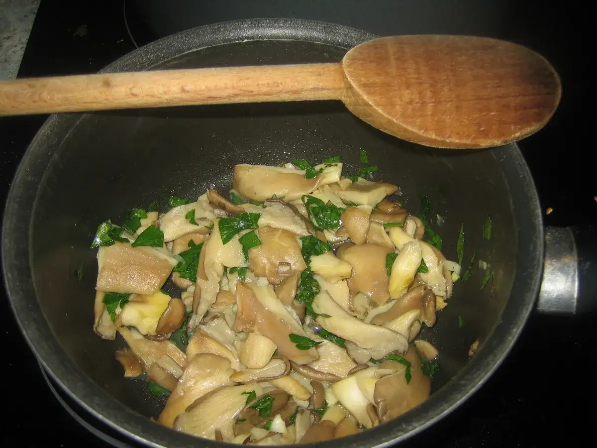 Poêlée de pleurotes frais, parsemée de persil, prête pour une délicieuse recette. Une cuillère en bois repose sur le bord.