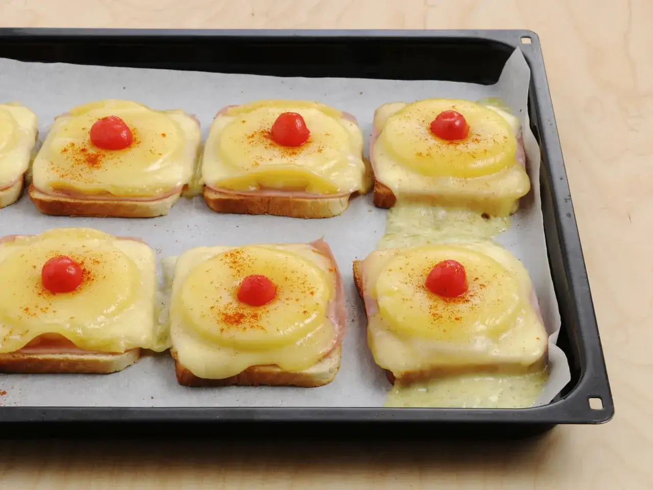 Sechs überbackene Toast Hawaii auf einem Backblech, mit geschmolzenem Käse, Ananasscheiben, Schinken und einer Kirsche obendrauf, bestreut mit Paprikapulver.