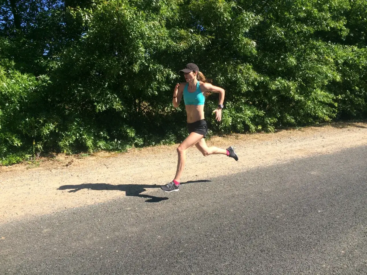Kvinna i bl&aring; sport-bh och svarta shorts springer p&aring; en grusv&auml;g. Hon har en keps och tr&auml;ningsskor. Solen lyser och skapar en l&aring;ng skugga. Hon verkar tr&auml;na f&ouml;r att springa 1 km tid.