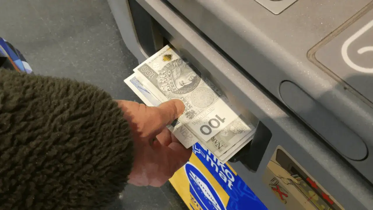 Ręka wkłada plik banknotów 100 zł do wpłatomatu. To szybka jednorazowa wpłata gotówki do banku.