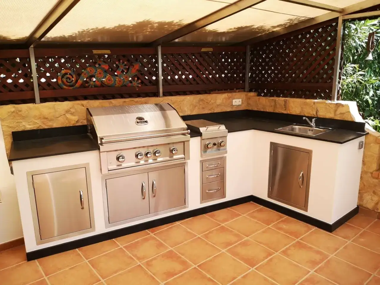 Porche con cocina exterior, con parrilla de acero inoxidable, fregadero y encimera negra. Decoración de lagarto en la celosía.