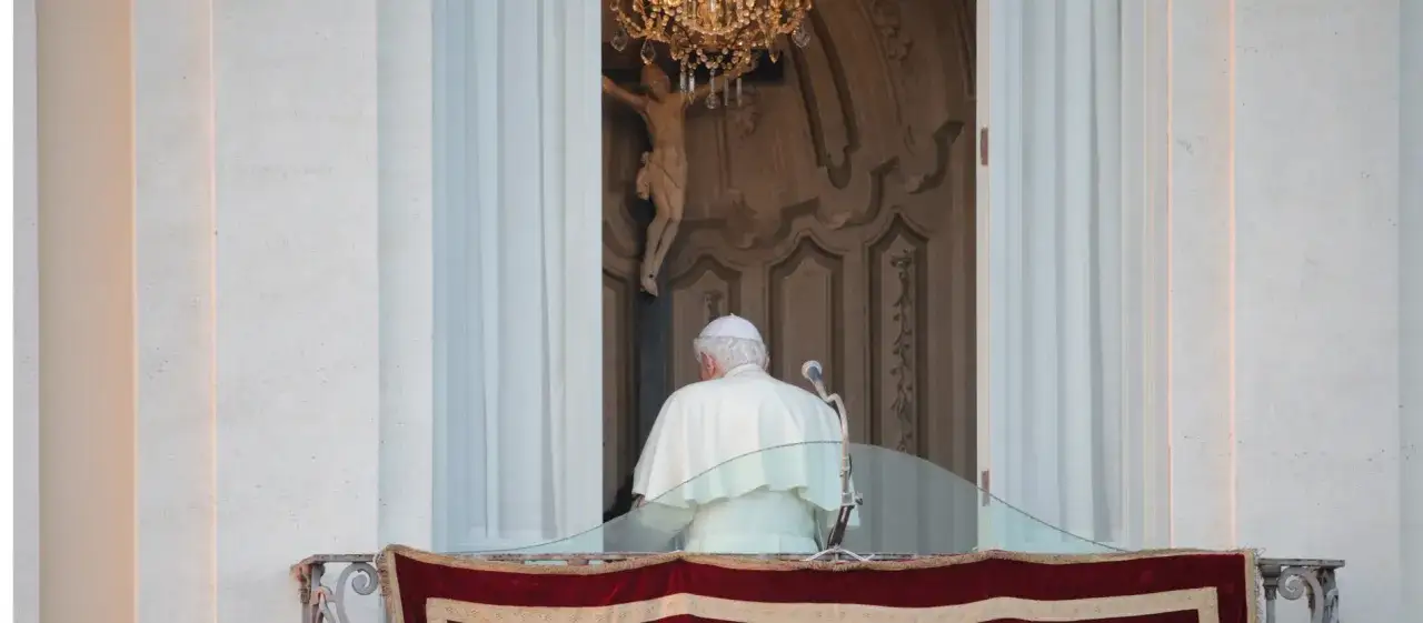 Der Papst steht auf dem Balkon, hinter ihm ein Kruzifix und ein Kronleuchter.