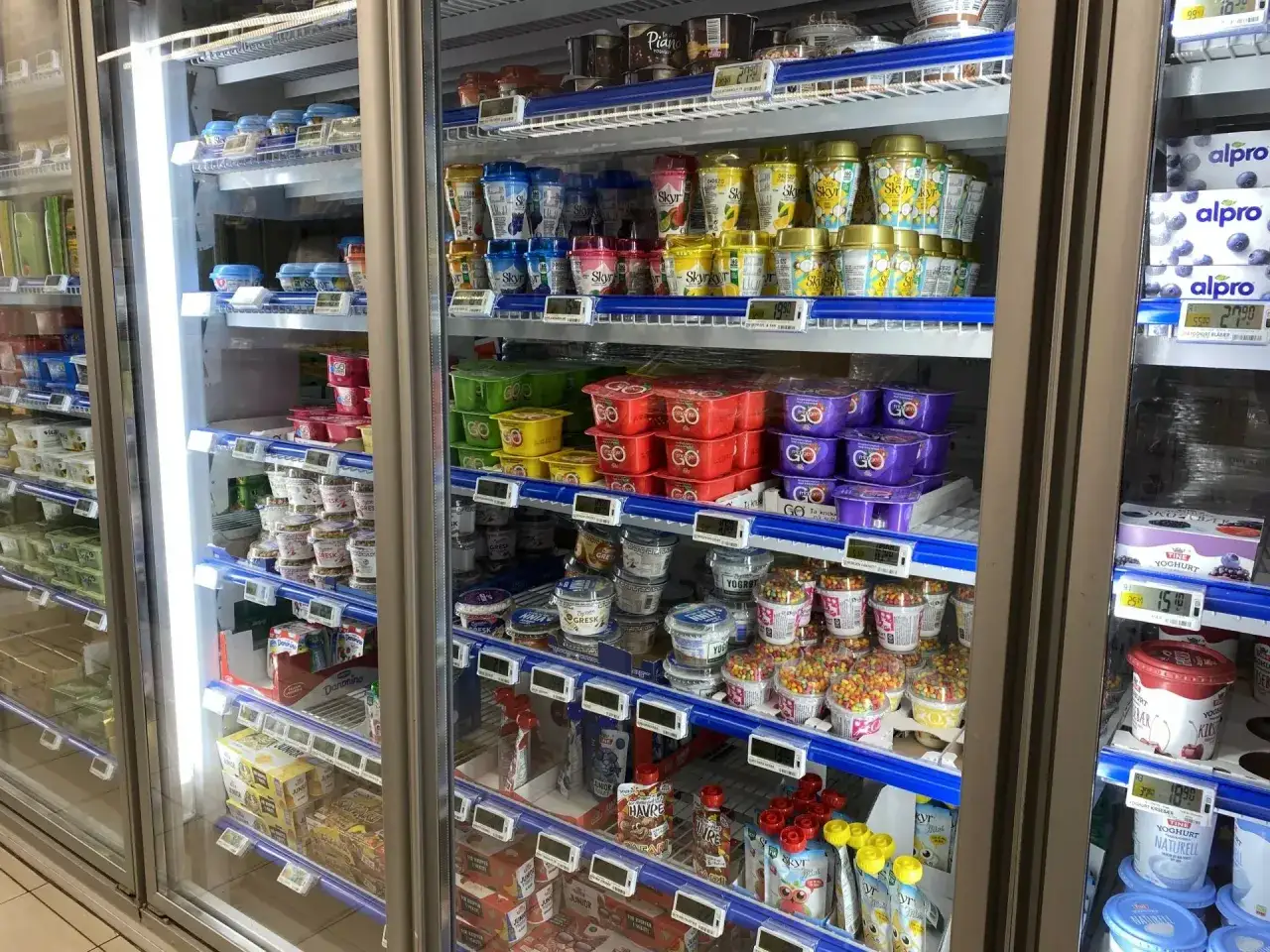 Lodówka z jogurtami i deserami. Widać różne marki i smaki, a także ceny, co daje pogląd na norwegia ceny w sklepach.