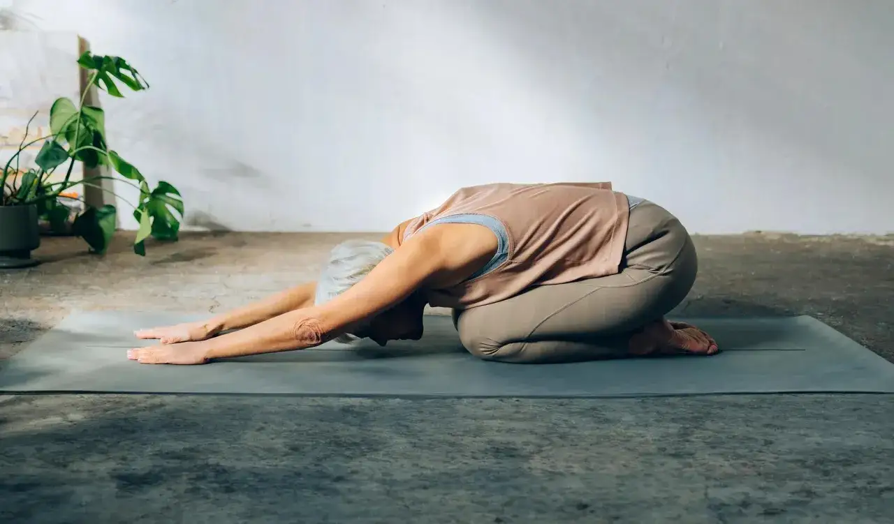&Auml;ldre kvinna i barnets position p&aring; yogamatta. Hennes armar str&auml;cks fram&aring;t, huvudet vilar mot golvet.