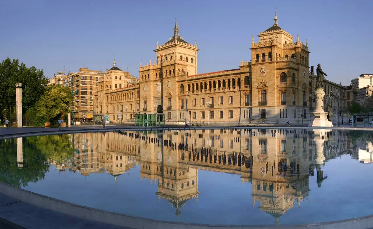 Le bâtiment principal de l'**universidad de Valladolid** se reflète dans un bassin d'eau, sous un ciel bleu clair.