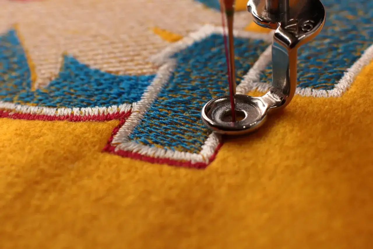 Gros plan sur une machine &agrave; coudre brodant un motif bleu et blanc sur un tissu jaune. Le fil rouge descend vers l'aiguille, pr&ecirc;t &agrave; &ecirc;tre int&eacute;gr&eacute; au fichier d'impression.