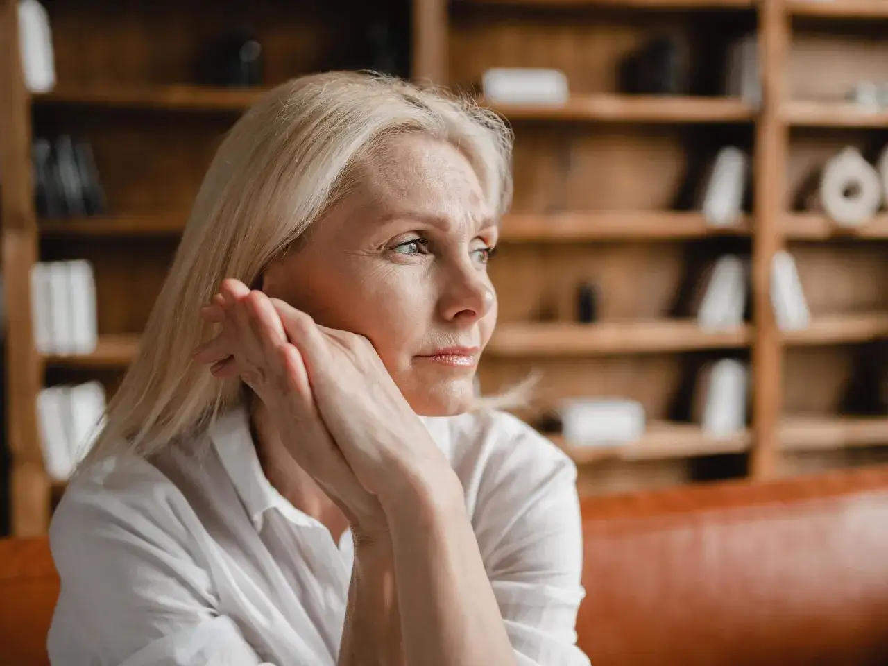 Nachdenkliche Frauen mittleren Alters mit blonden Haaren, die auf einem braunen Sofa sitzen, mit Bücherregalen im Hintergrund.
