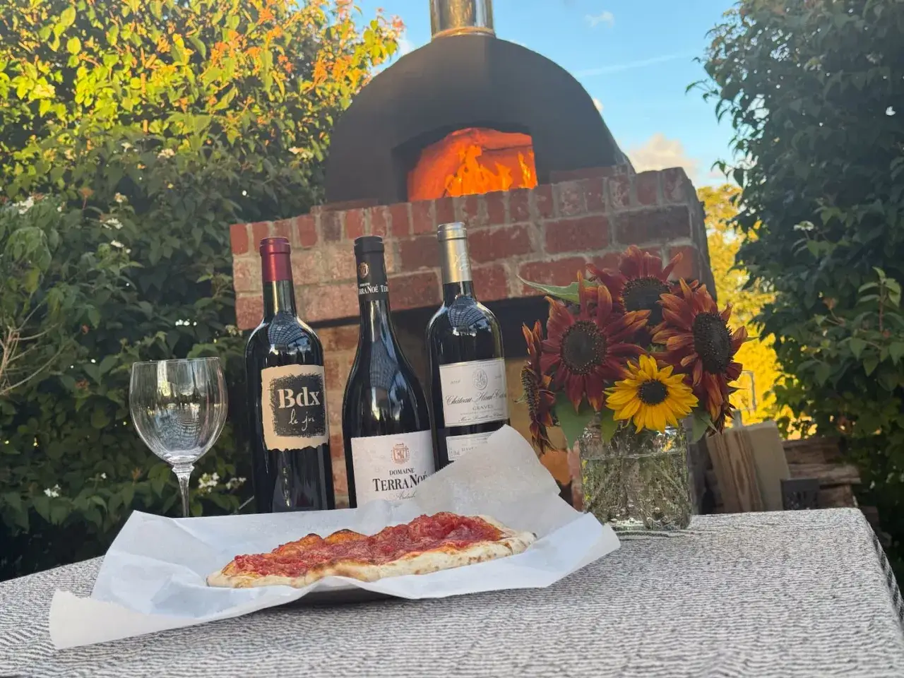 Welcher Wein zu Pizza? Drei Rotweinflaschen, ein Glas, eine Pizza und Sonnenblumen vor einem Pizzaofen.