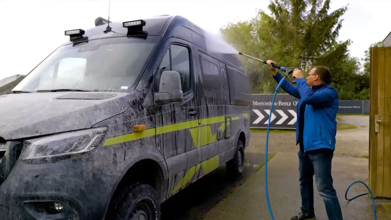 Un homme nettoie un camping-car increvable, couvert de boue, avec un nettoyeur haute pression.