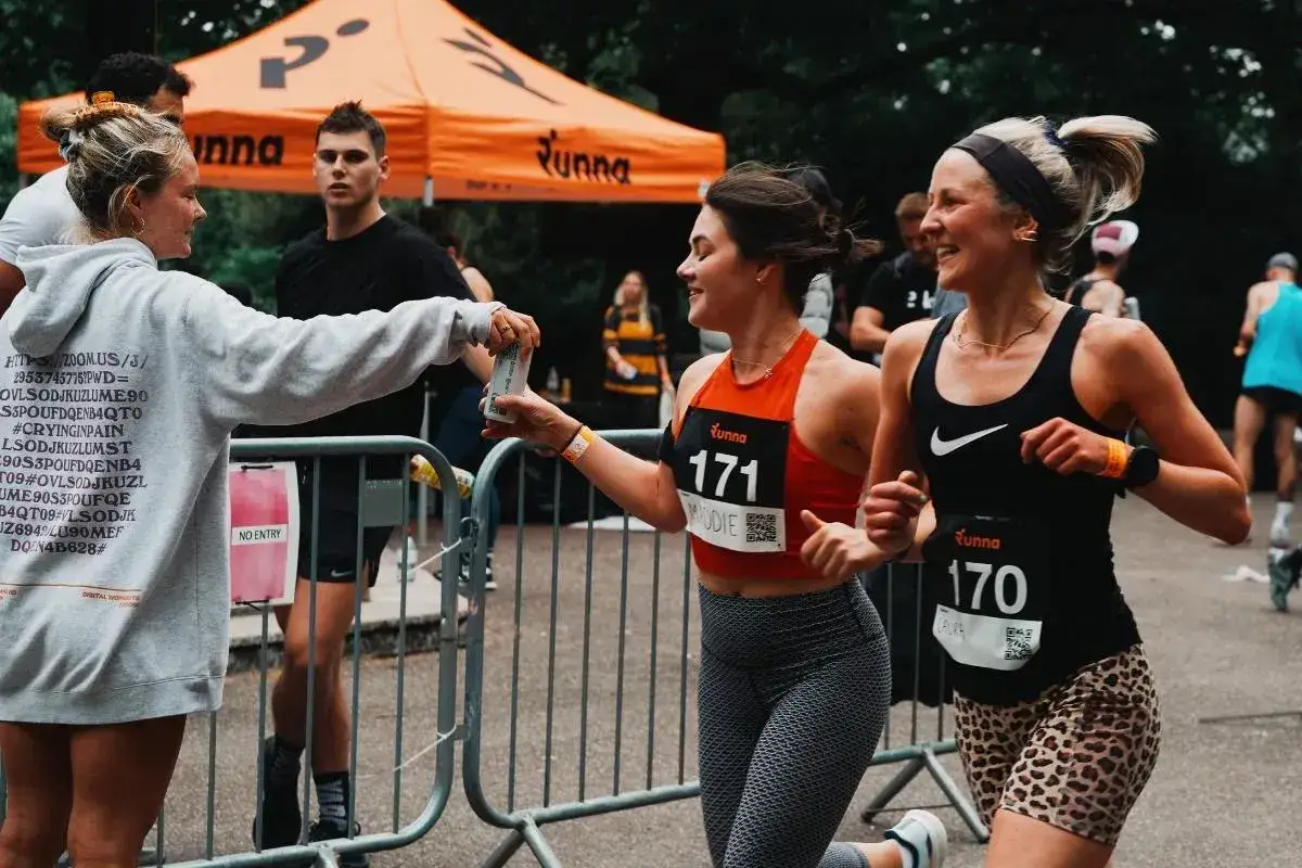 L&auml;uferinnen erhalten nach dem Marathon Erfrischungen. Eine Frau im grauen Hoodie reicht eine Dose, w&auml;hrend zwei L&auml;uferinnen mit Startnummern vorbeilaufen.