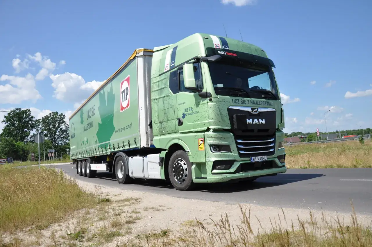 Zielony zespół pojazdów MAN z naczepą TIP jedzie po drodze. Na naczepie widnieje napis