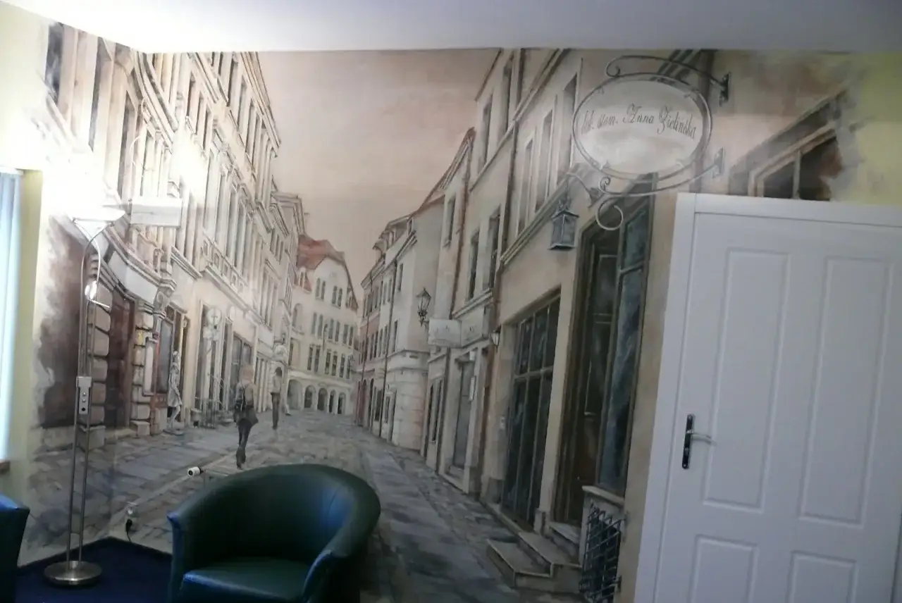 Malarstwo artystyczne wnętrz: iluzja uliczki starego miasta na ścianie. Wygodny fotel i lampa dopełniają scenografię.