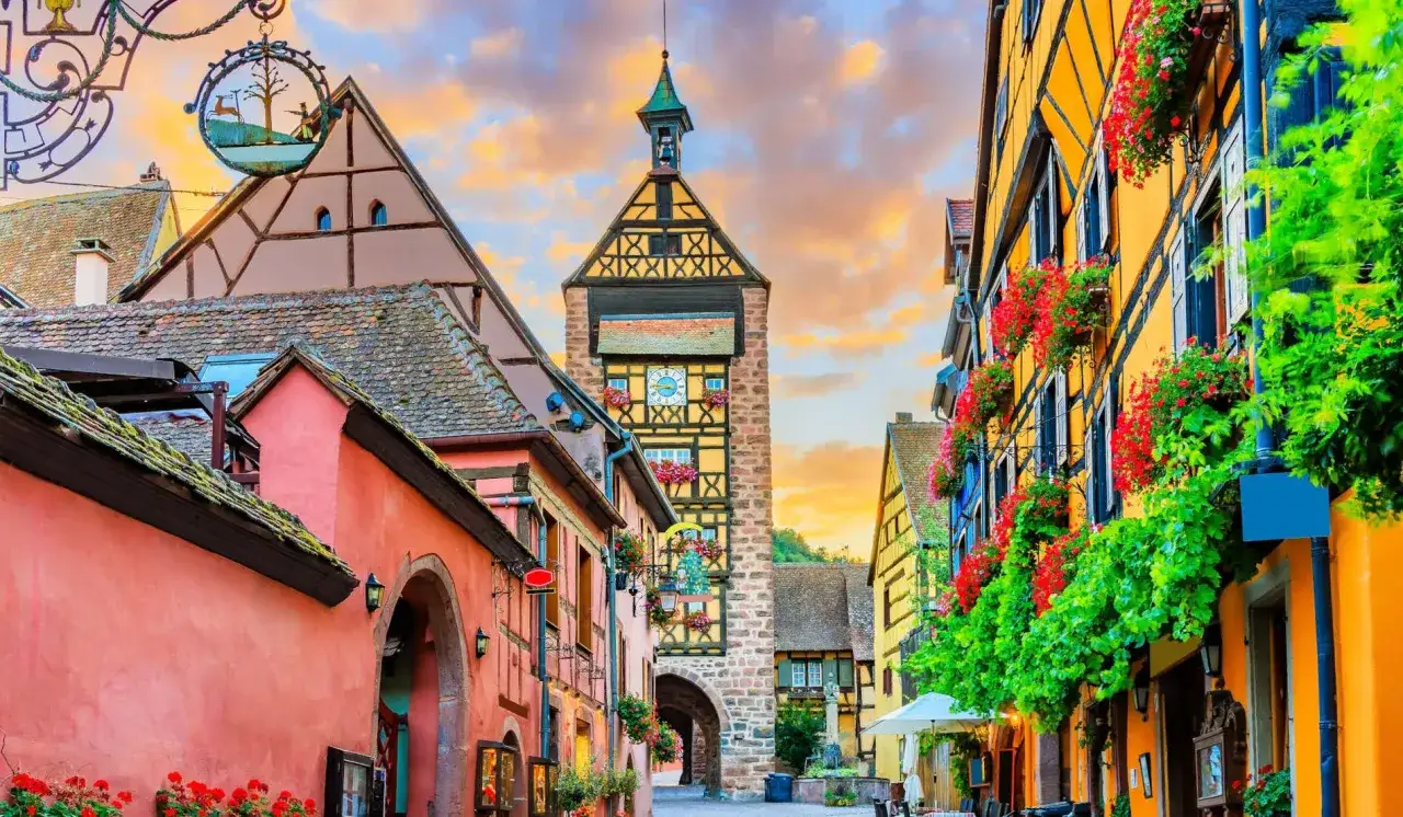 Calle pintoresca con casas de colores y flores, ideal que ver en Alsacia. Torre con reloj y cielo al atardecer.