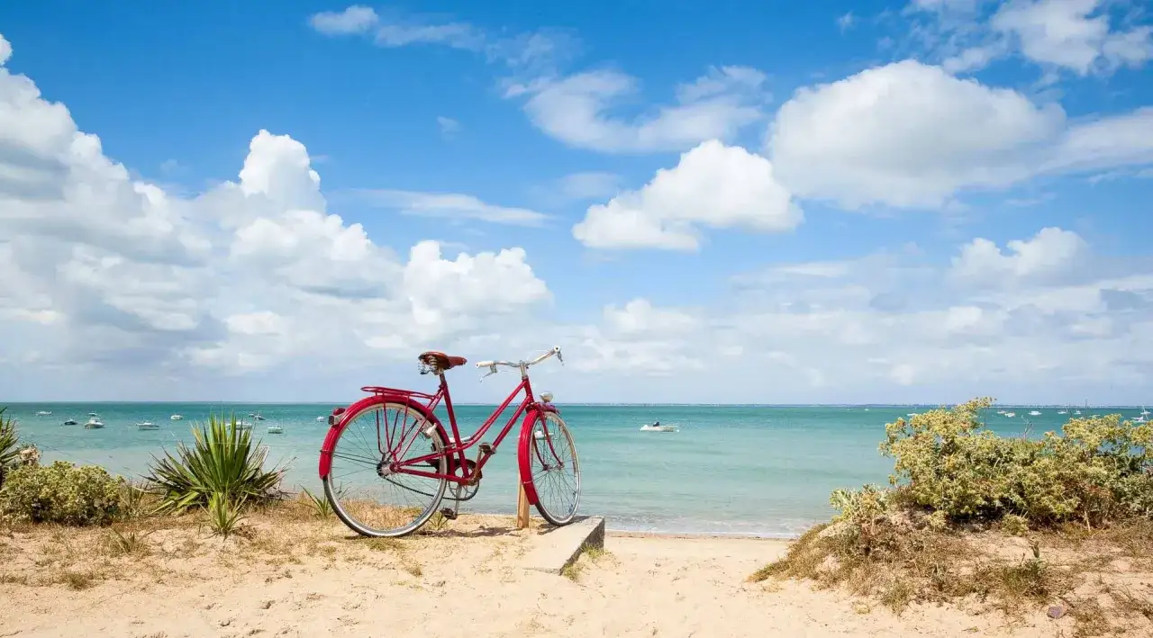 Vélo rouge sur une plage de sable fin, avec la mer turquoise et des nuages blancs à l'horizon. Une invitation à explorer l'île de Ré.