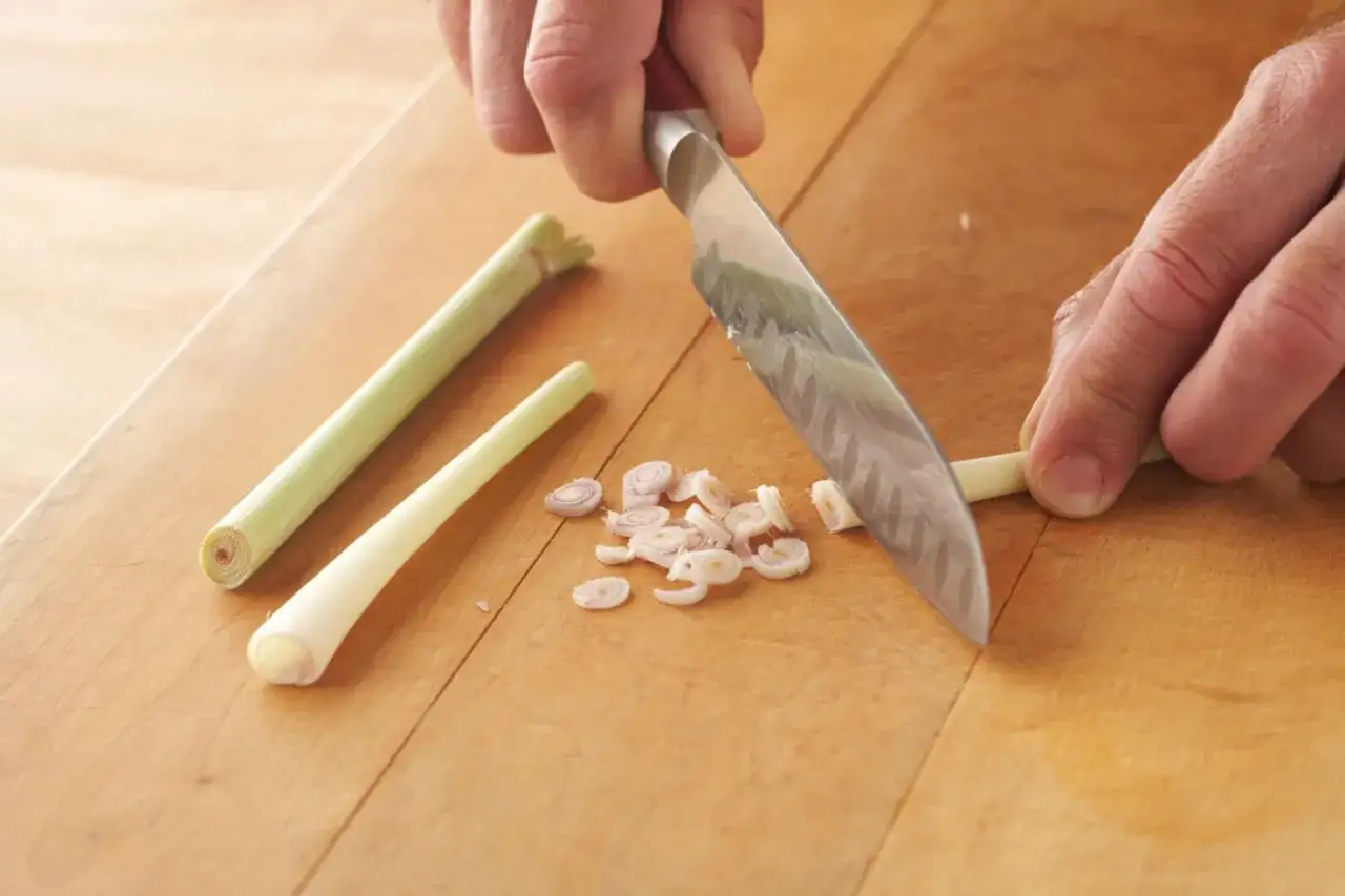 Hands chop fresh lemongrass on a wooden cutting board, creating a pile of finely sliced pieces.