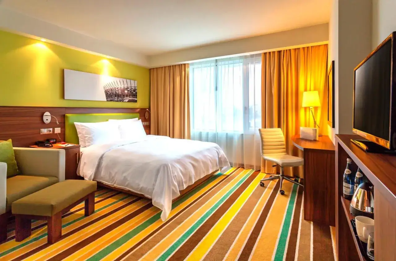 Pokój w Hampton by Hilton Warsaw: łóżko, kanapa, biurko, TV i kolorowy dywan.