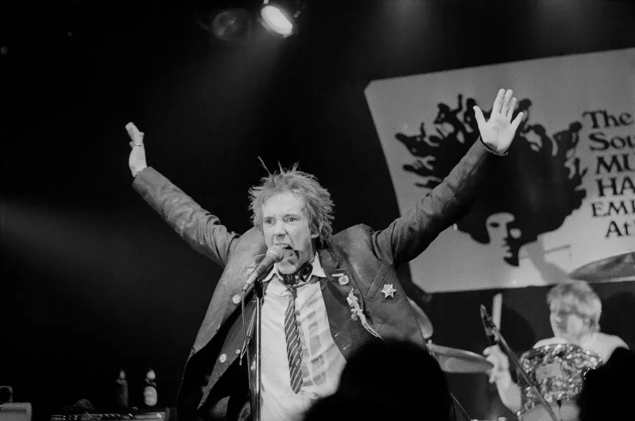 Le chanteur des Sex Pistols, Johnny Rotten, hurle au micro, bras lev&eacute;s, sur sc&egrave;ne.