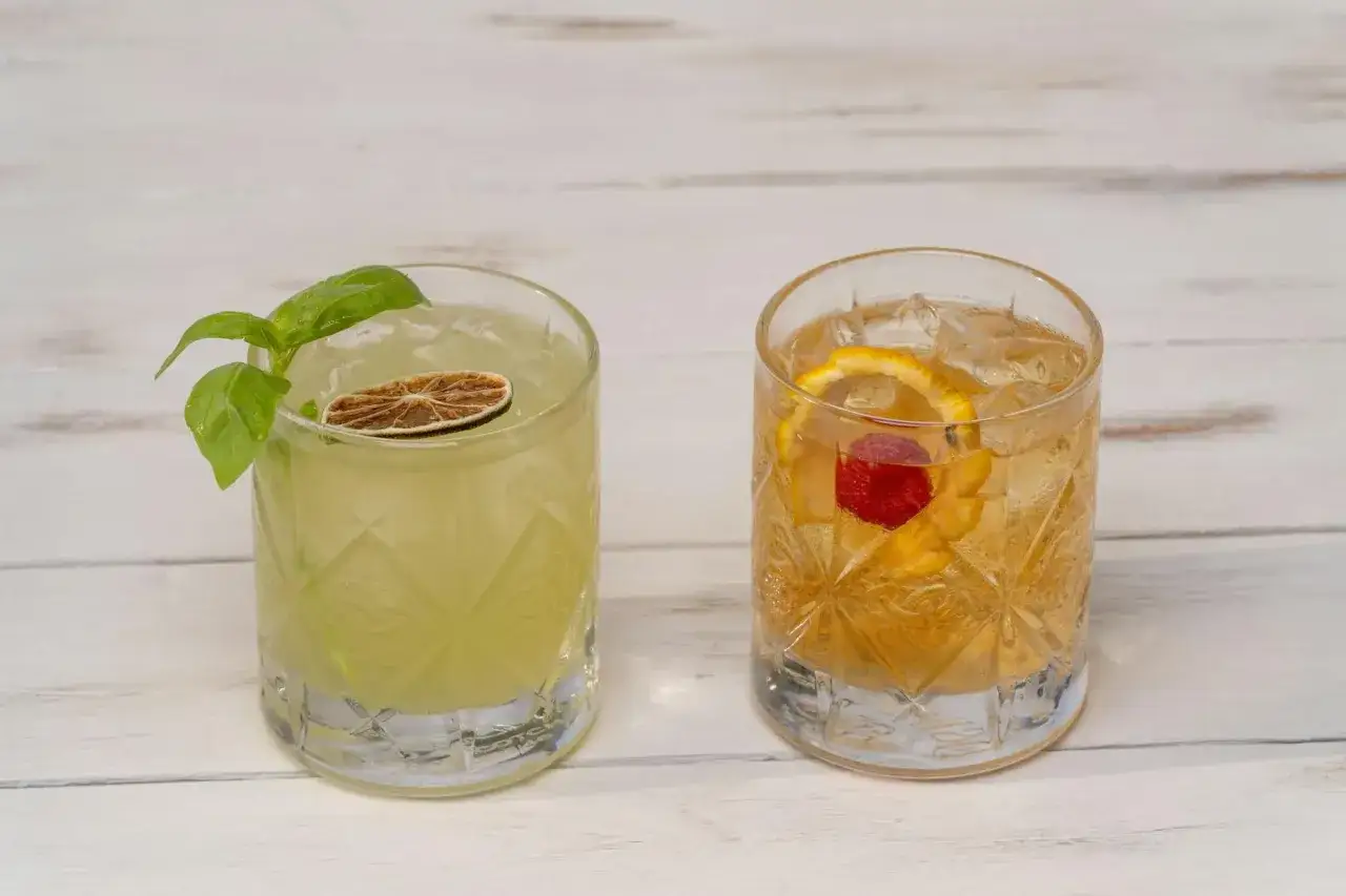Dwa drinki w szklankach: jeden zielony z plasterkiem limonki i bazylią, drugi bursztynowy z pomarańczą i maliną. Idealne do eksperymentów, jak zrobić bitters domowej roboty z piołunu i korzenia arcydzięgla.