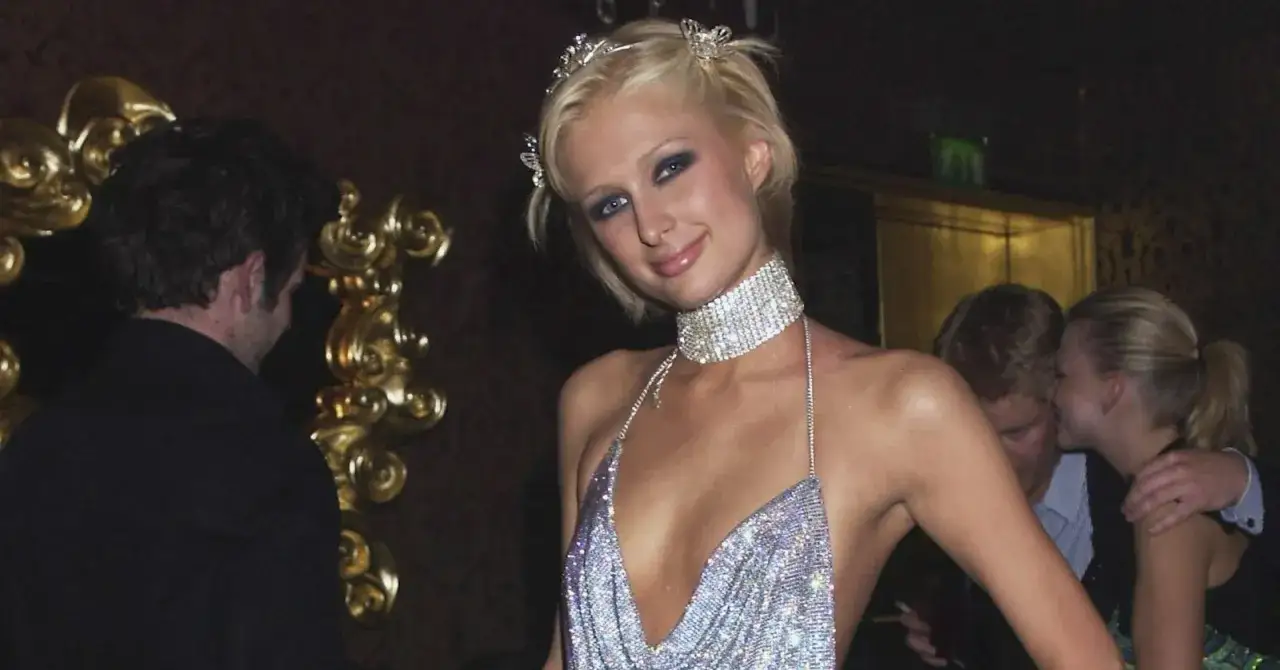 Paris Hilton gl&auml;nzt in einem funkelnden 2000er Mottoparty-Outfit mit Diadem und Choker. Im Hintergrund tuscheln andere G&auml;ste.