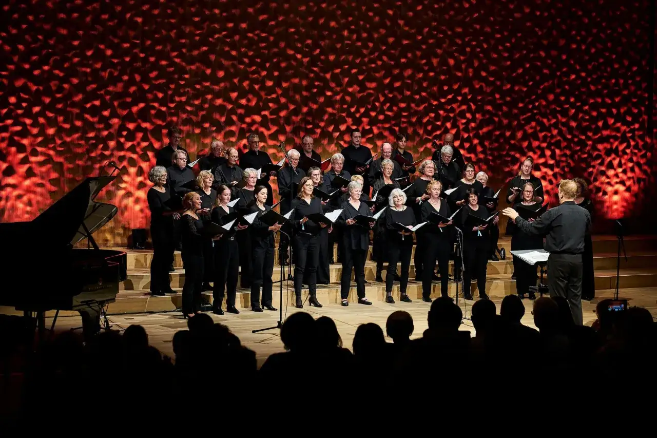 Chor singt vor rotem, gemustertem Hintergrund. Ein Klavier steht links. Die Aufführung könnte Teil des Kirchentag 2029 sein.