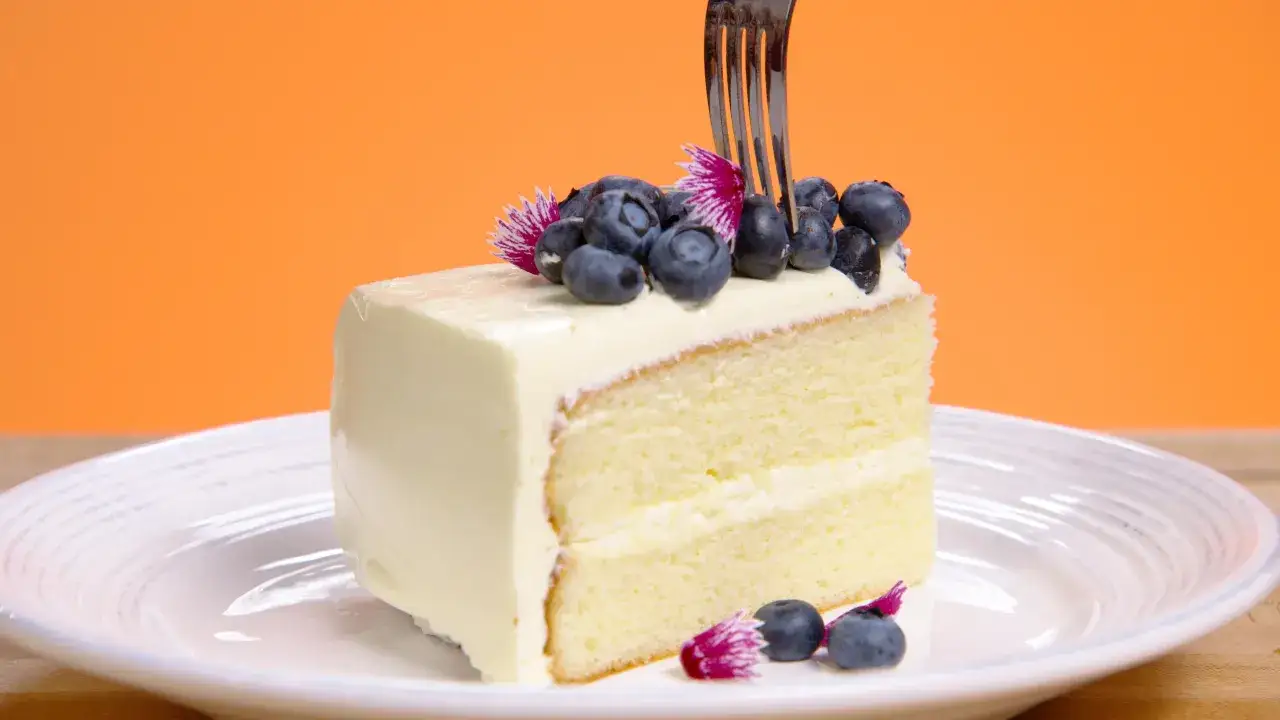 Tranche de gâteau moelleux, recouverte d'une onctueuse **butter cream** et garnie de myrtilles fraîches. Une fourchette s'apprête à déguster ce délice.