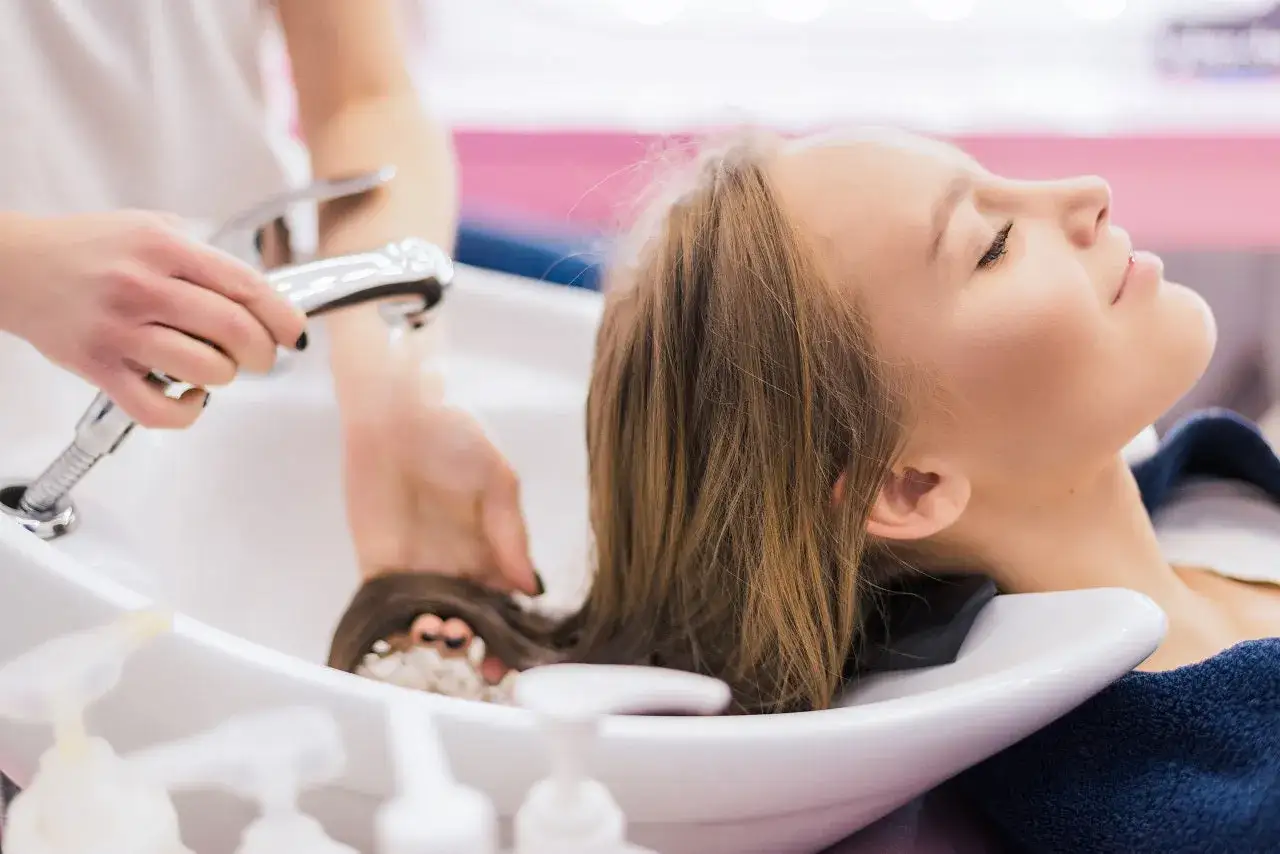 Eine Frau genie&szlig;t eine entspannende Haarw&auml;sche. Die Friseurin sp&uuml;lt ihre langen, braunen Haare mit Wasser ab. Sie hat ihre Augen geschlossen und l&auml;chelt sanft. Die Frau l&auml;sst ihre Haare nur mit Conditioner waschen.