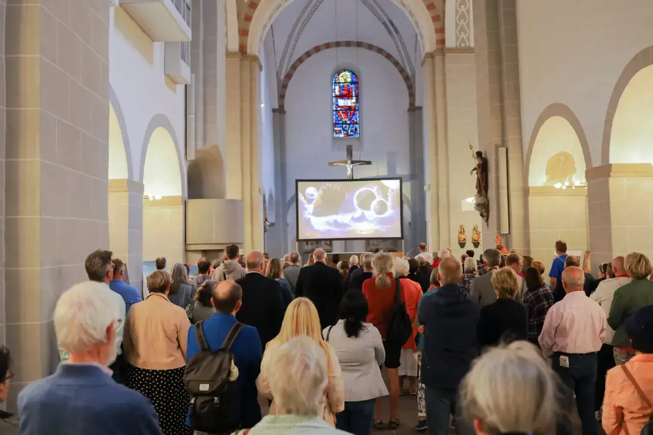 Menschen versammeln sich in einer Kirche, um eine Projektion zu sehen. Ein Bild von Planeten wird auf eine Leinwand projiziert, w&auml;hrend eine Figur, die an den **Papst ki** erinnert, auf einer Statue steht.