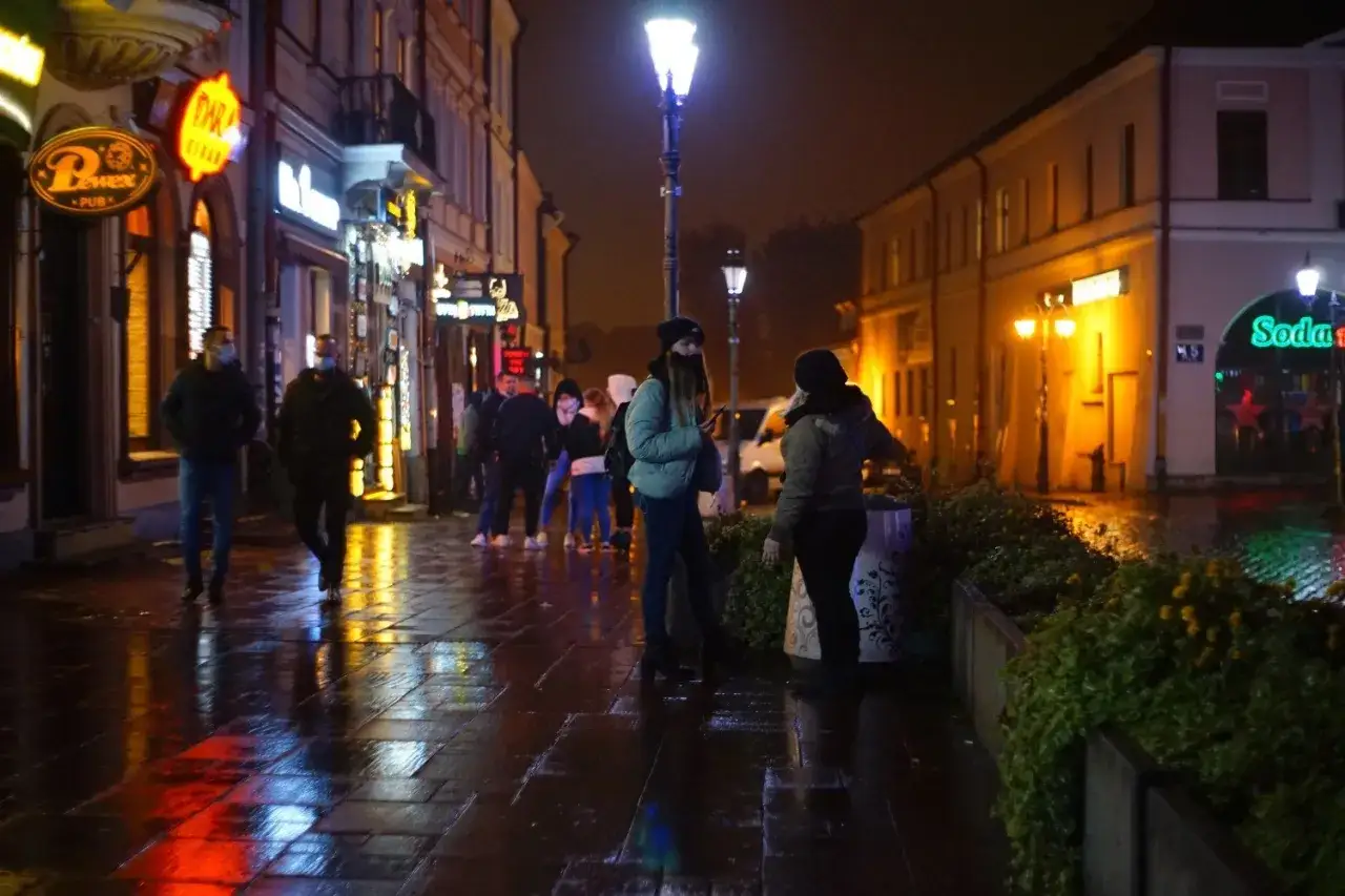 Mokry rzeszów rynek nocą. Ludzie w maskach spacerują po oświetlonej ulicy, odbijając się w mokrej nawierzchni.