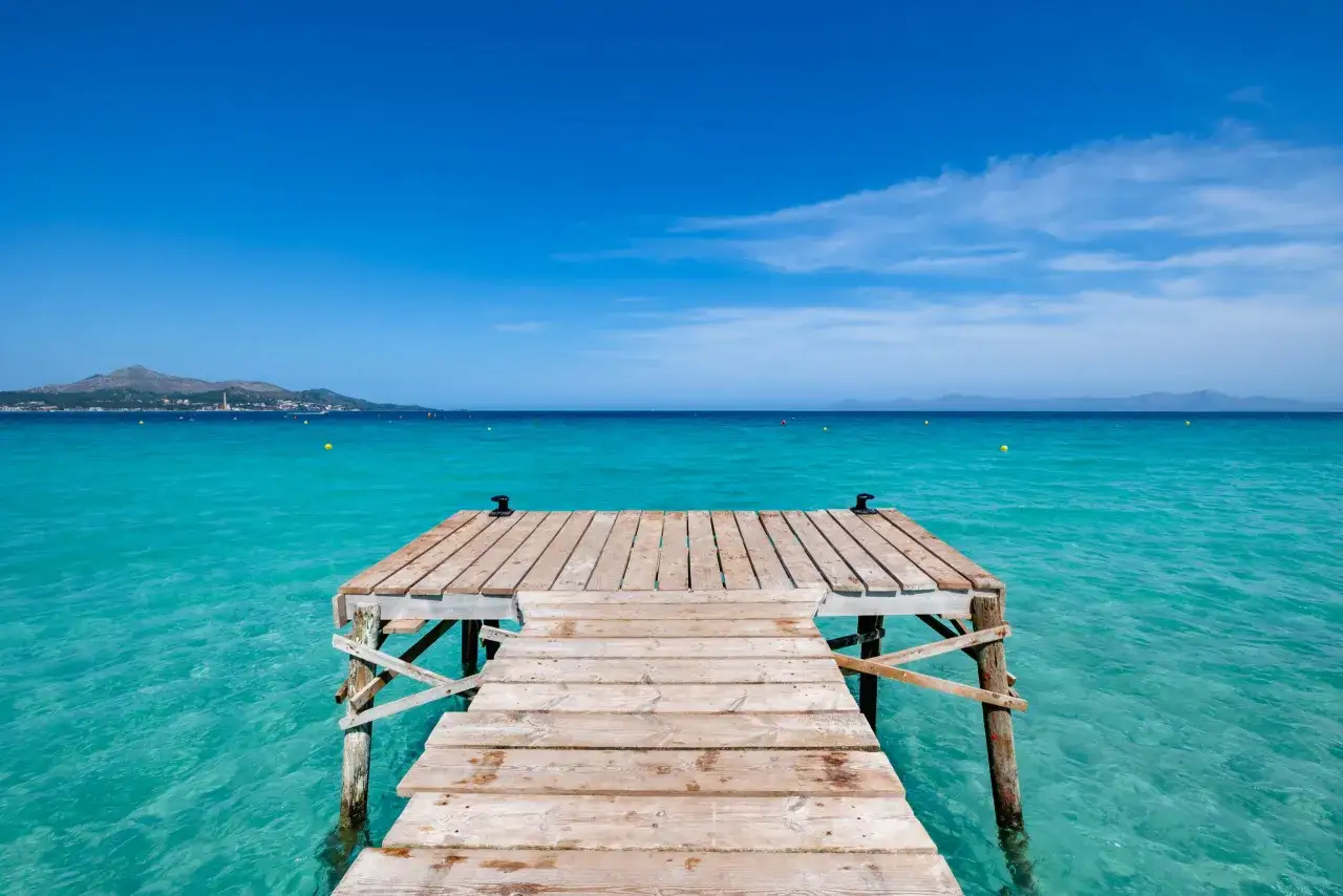 Hol dir die besten Mallorca Tipps für deinen Traumurlaub: Ein Holzsteg führt ins kristallklare, türkisfarbene Meer vor einer Bergkulisse.