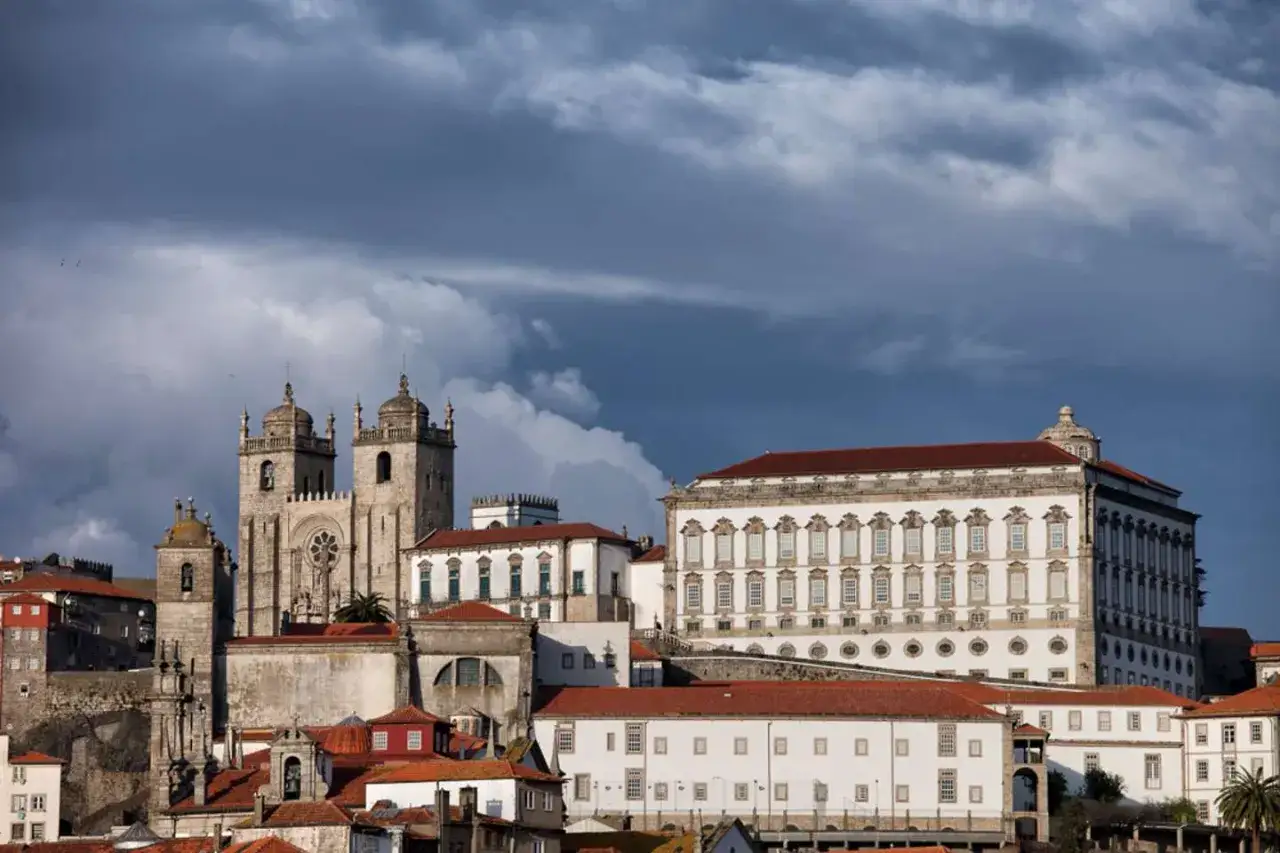 Edificios históricos de Oporto bajo un cielo amenazante, con la catedral y el palacio a la vista. El **oporto con lluvia** se intuye en las nubes oscuras.