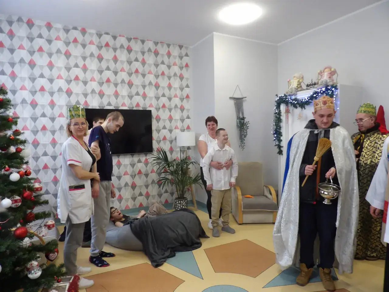 Grupa osób w strojach przypominających Trzech Króli przygotowuje się do kolędy. W tle choinka i świąteczne dekoracje.