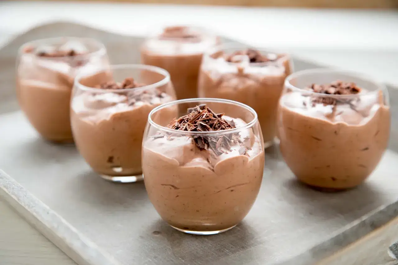 Seis copas de mousse de chocolate de la abuela, cubiertas con virutas de chocolate, dispuestas en una bandeja de madera.