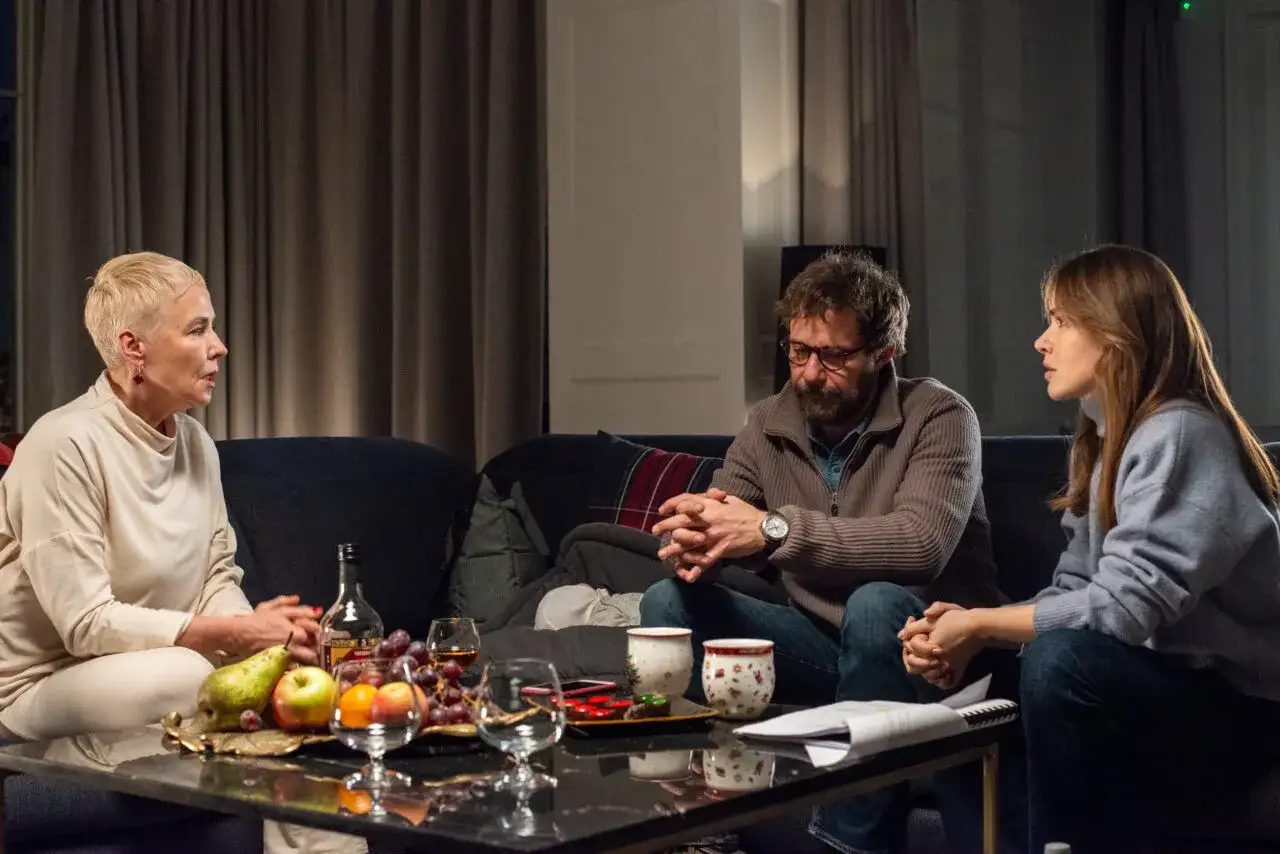 Trzy osoby siedzą na kanapie, rozmawiając przy stoliku z owocami i alkoholem. Czy to początek zabawnych komedii?