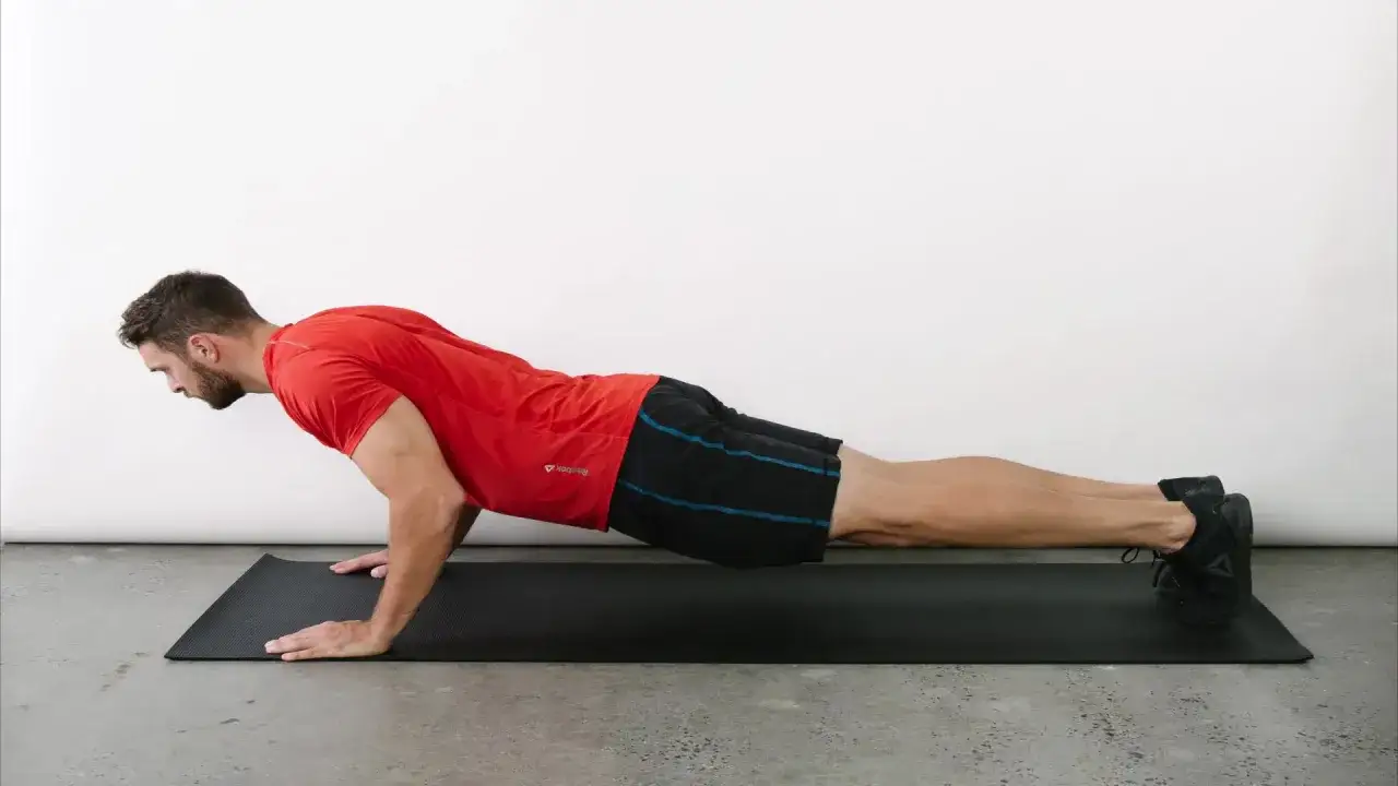 Un homme en t-shirt rouge et short noir est en position de planche sur un tapis de yoga, prêt à apprendre comment faire des pompes.