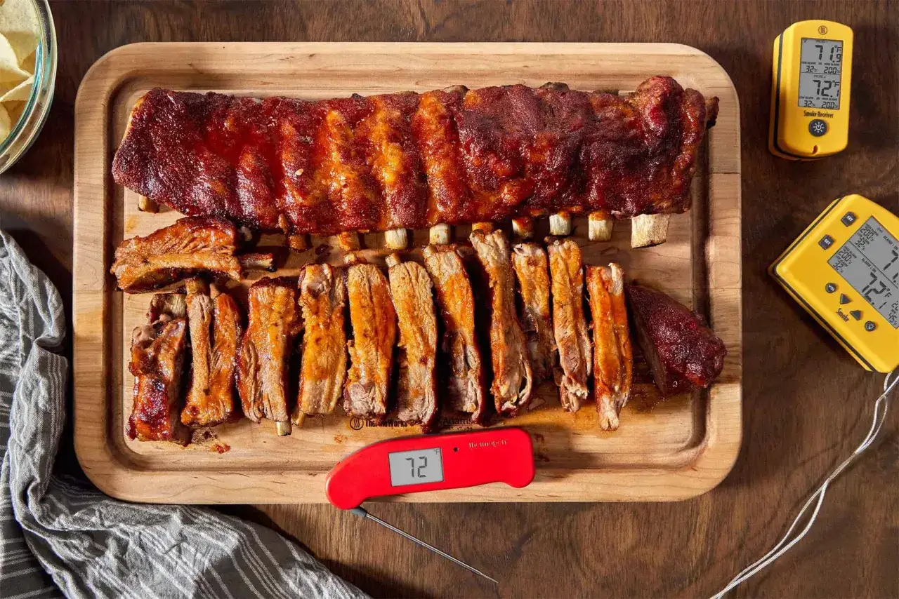 Un plat de ribs de porc grillés, nappés de sauce barbecue, sur une planche en bois. Deux thermomètres de cuisson sont visibles.