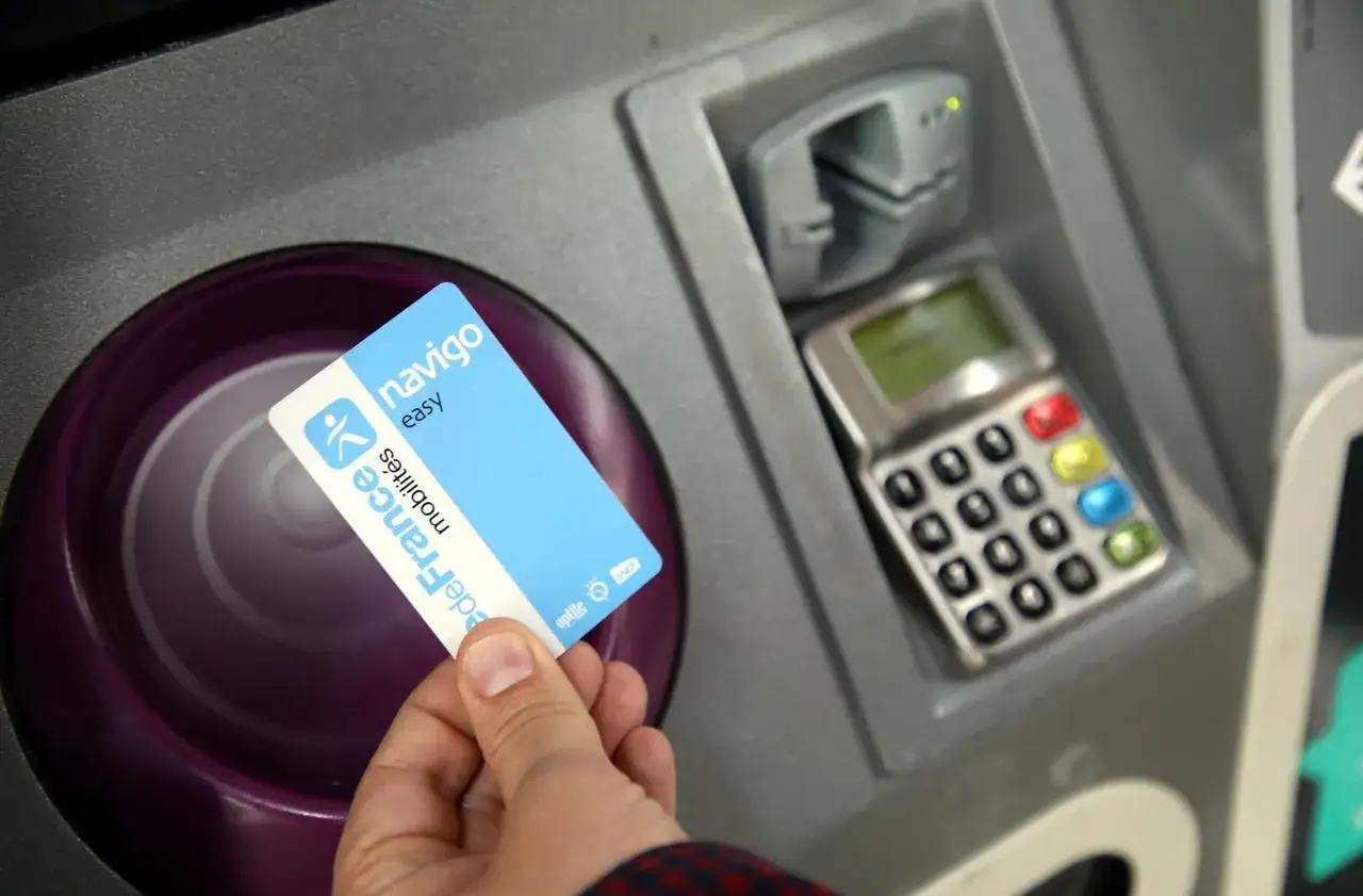 Main d'une personne tenant une carte Navigo Easy, un titre de transport pour le métro parisien, près d'une borne.