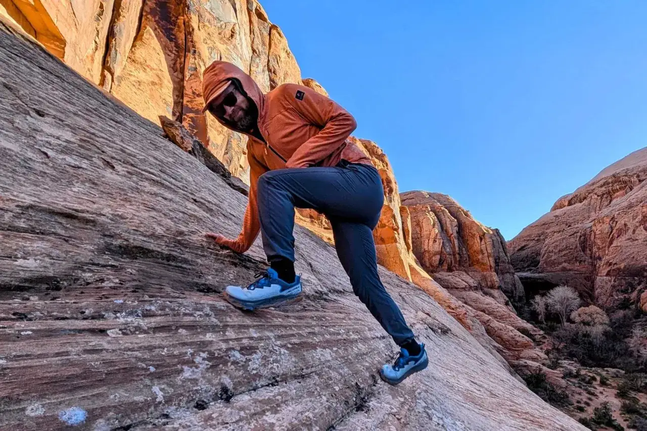 Man in orange hoodie and Altra timp 6 shoes climbs a rocky desert slope under a clear blue sky.
