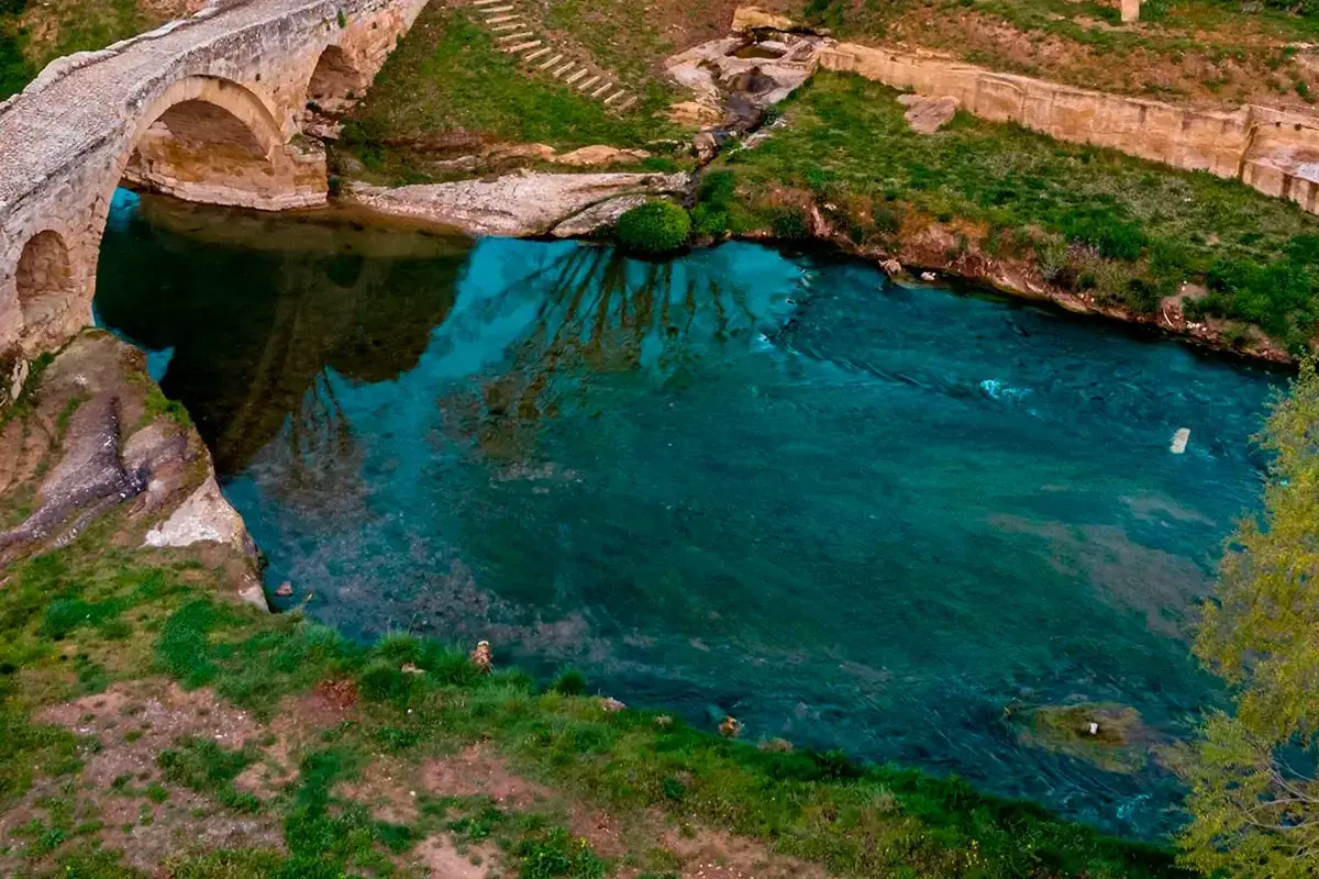 Un puente de piedra cruza un río de aguas turquesas, creando piscinas naturales en La Rioja. Reflejos de árboles y vegetación adornan la escena.