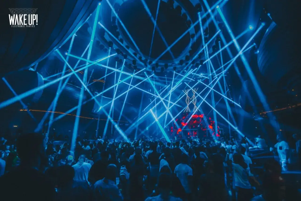 Una multitud baila bajo un espectáculo de luces azules en un evento de música electrónica. El logo