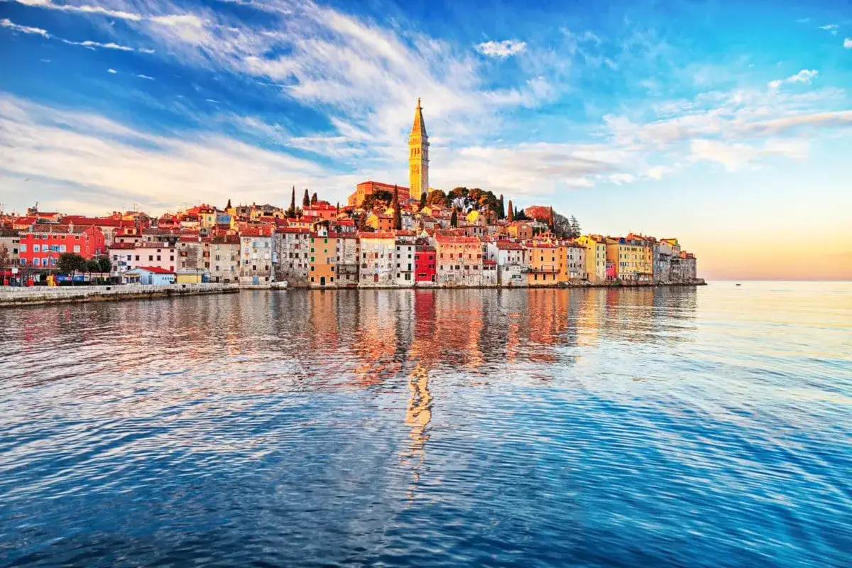 Bunte Häuserfront am Wasser in Rovinj, Kroatien. Der Kirchturm ragt über die Dächer, während sich die Stadt im ruhigen Meer spiegelt.