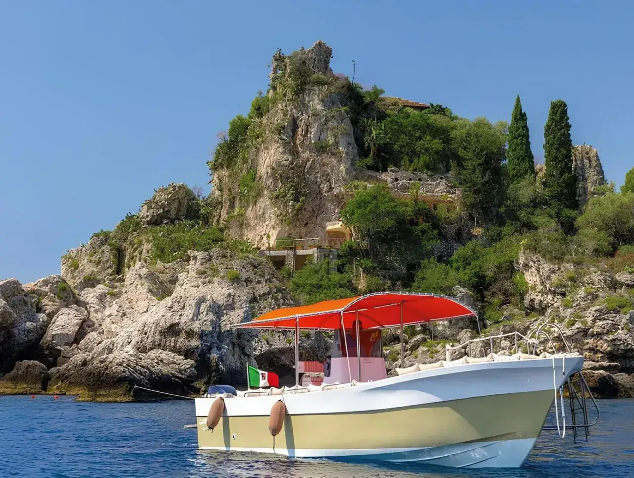 Barca con bandiera italiana vicino alla costa rocciosa, con vegetazione lussureggiante e un sentiero che porta verso l'alto. Un'immagine che evoca la bellezza della grotta azzurra Taormina.