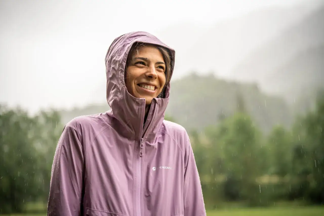 L&auml;chelnde Frau im Regen, gesch&uuml;tzt durch eine wasserdichte 5000 mm Jacke. Tropfen perlen ab, w&auml;hrend sie die Natur genie&szlig;t.