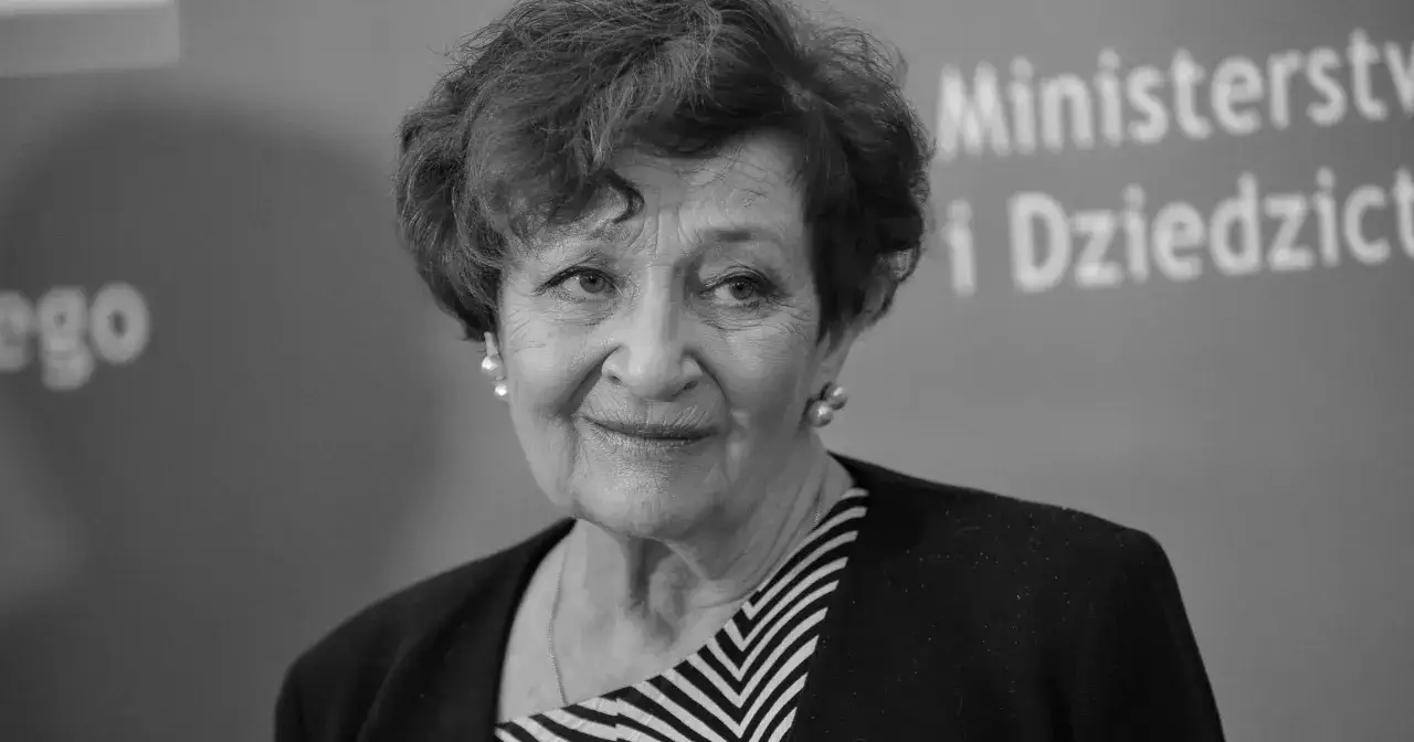 Uśmiechnięta Ewa Dałkowska, z perłowymi kolczykami, na tle napisu