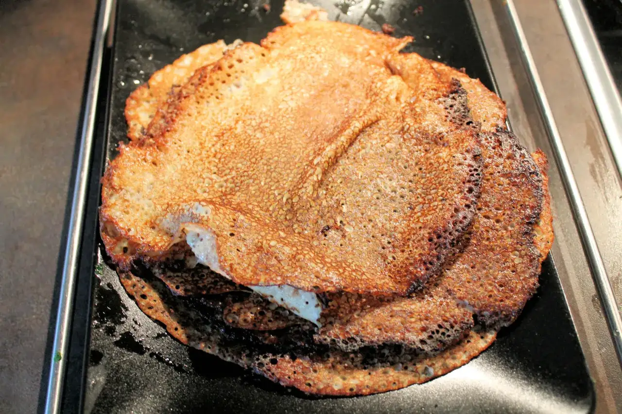 En hög med gyllene plättar pannkakor, perfekt stekta med små bubblor på ytan, ligger på en svart platta.