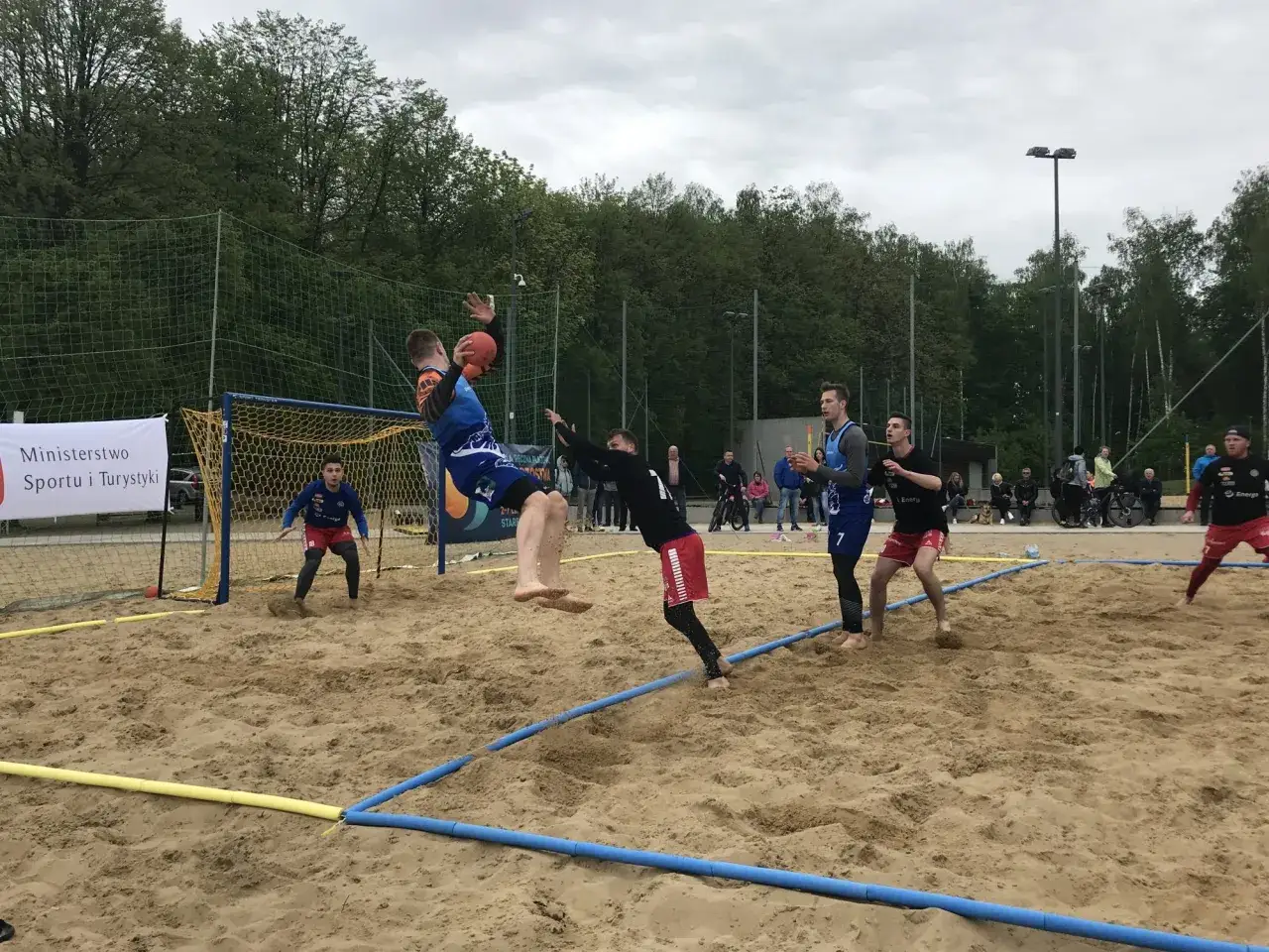 Zawodnicy walczą o piłkę podczas meczu piłki ręcznej plażowej. Jeden z graczy wyskakuje do rzutu, podczas gdy inni pr&oacute;bują go zatrzymać.