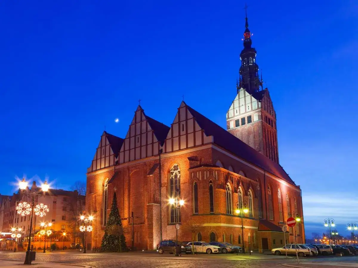 Wieczorny widok na stare miasto Elbląg. Imponująca ceglana katedra z wysoką wieżą, ozdobiona świątecznymi iluminacjami, góruje nad placem.