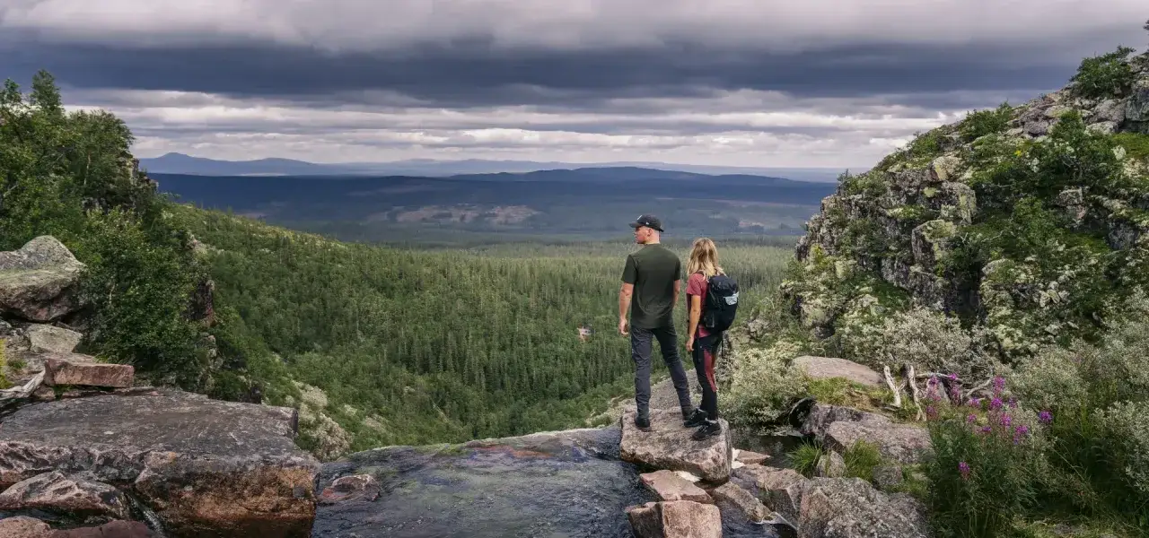 Två vandrare njuter av utsikten över en vidsträckt skog och berg i Dalarna. Perfekt för att utforska vandringsleder Dalarna.