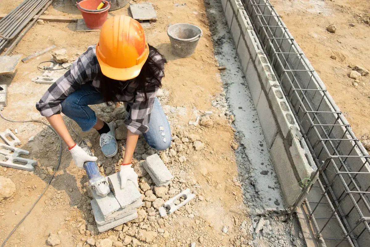 Budowlaniec w kasku obrabia bloczki betonowe, przygotowując je do budowy fundamentu z betonu c12/15.