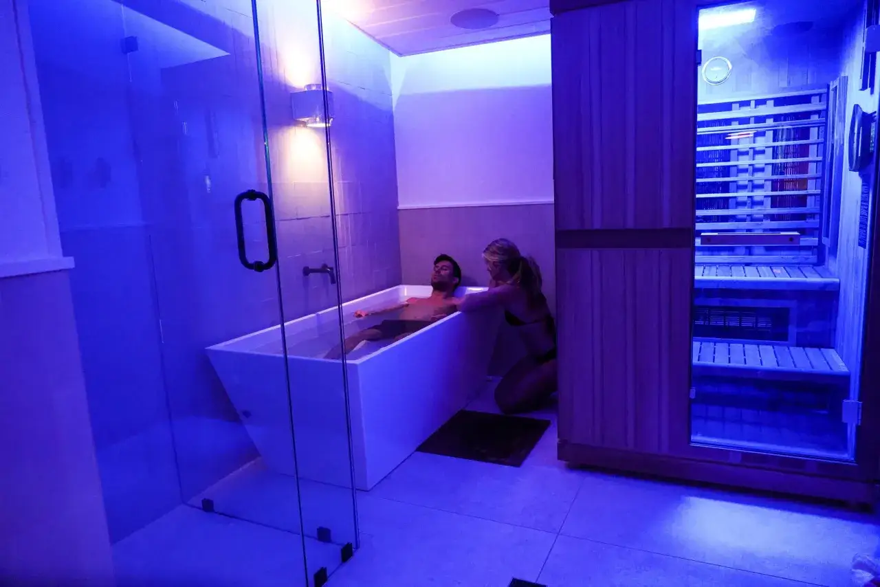 Coppia in un bagno moderno con vasca e sauna. Lei accarezza lui nella vasca, un momento di relax che ricorda la **doccia svedese**.